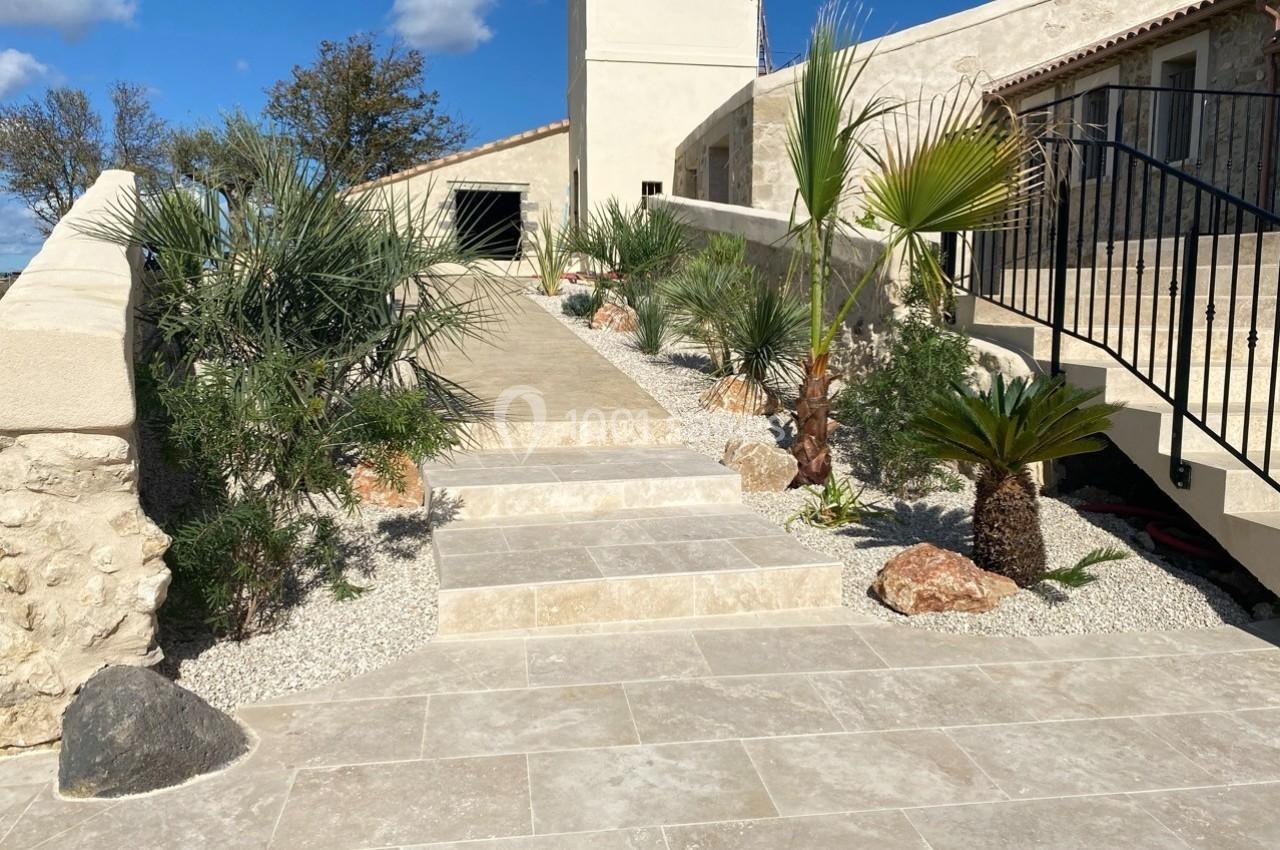 Escalier en pierre entouré de plantes méditerranéennes, menant à une maison en pierre sous un ciel bleu.