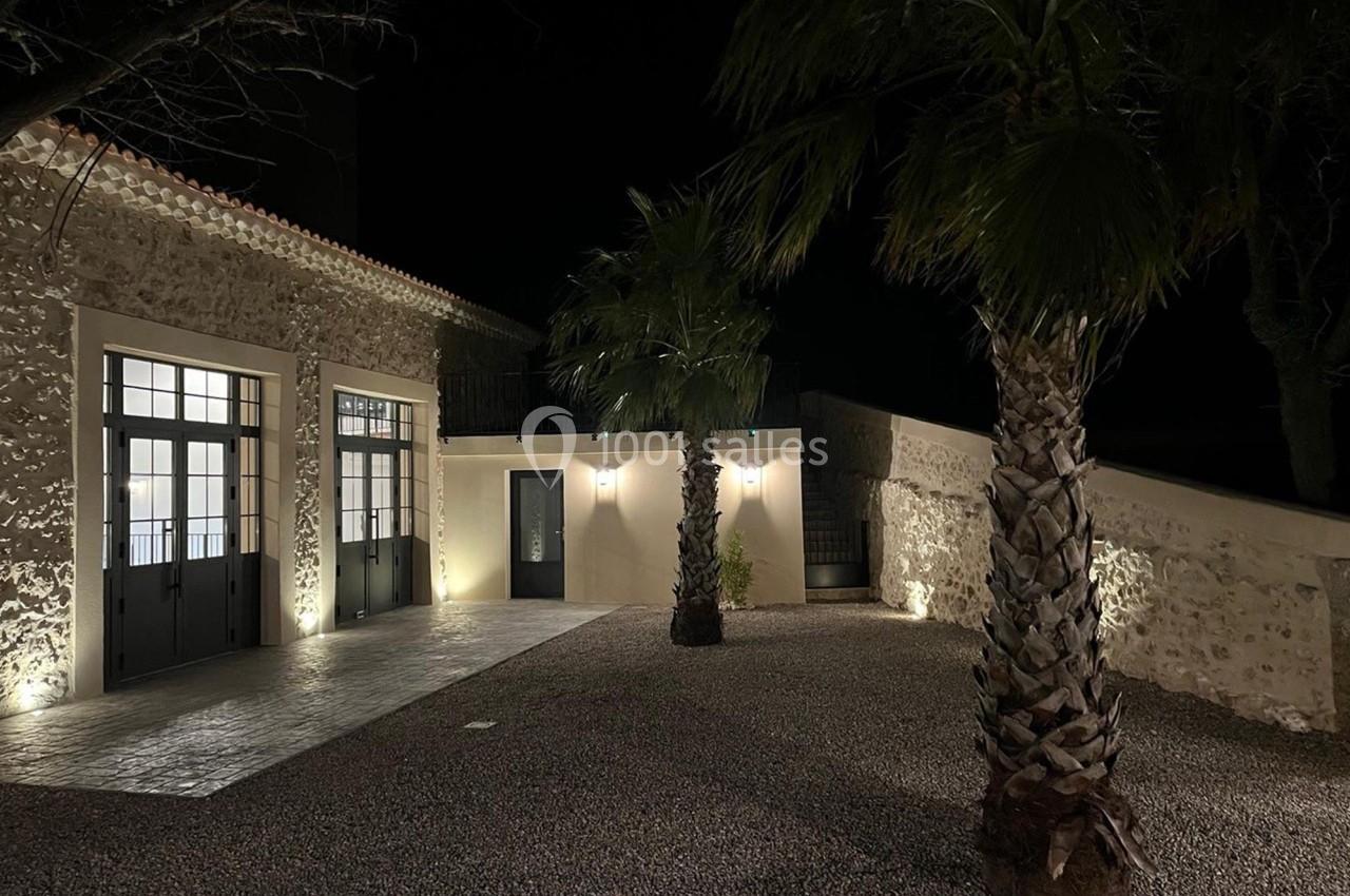 Cour extérieure éclairée de nuit, avec façade en pierre, grandes portes vitrées et palmiers sur sol gravillonné.