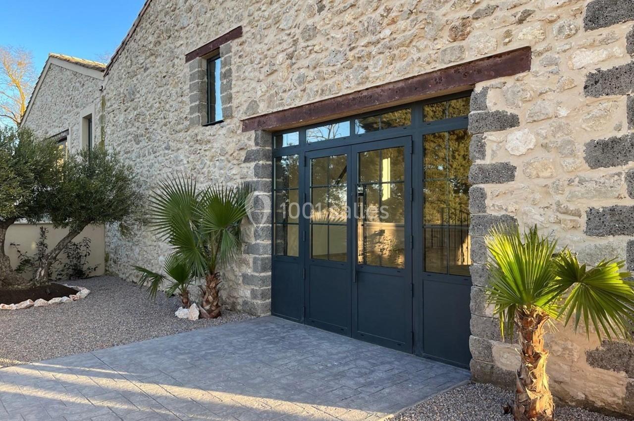 Façade en pierre avec une grande porte vitrée noire, entourée de palmiers et d'un sol en gravier.