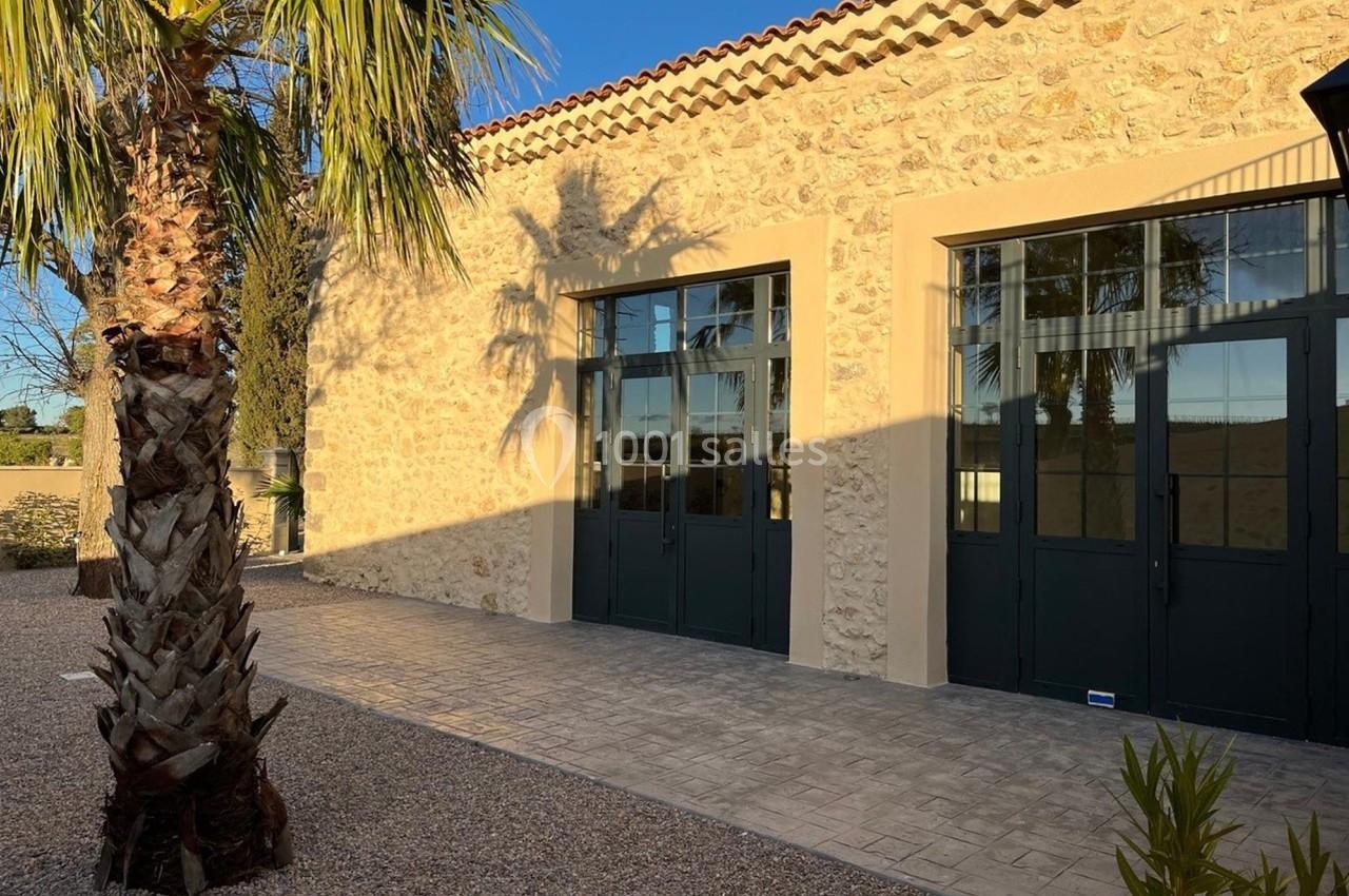 Façade en pierre d'un bâtiment avec grandes portes vitrées, éclairée par le soleil, entourée de végétation.