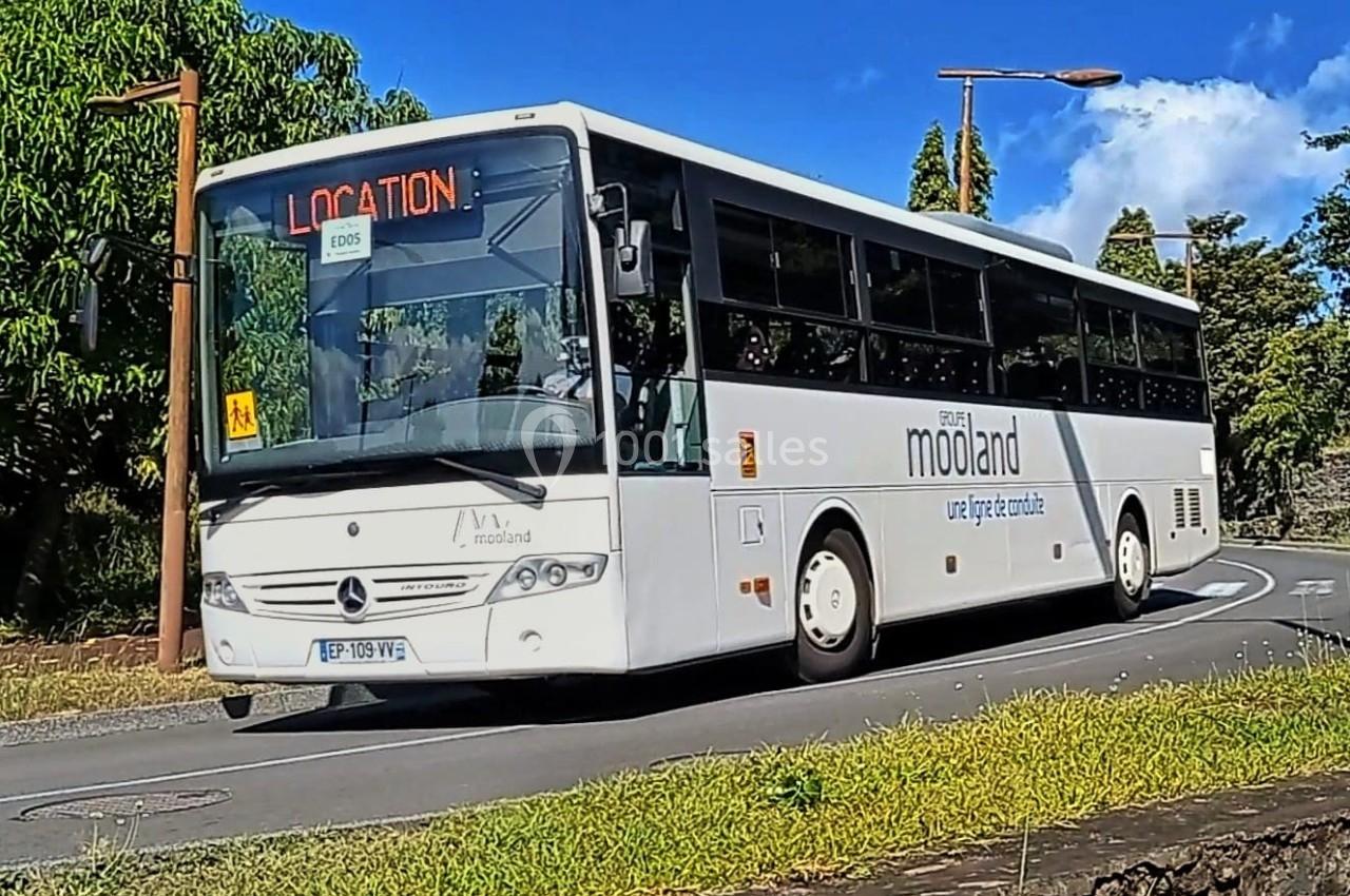 Autocar blanc circulant sur une route bordée de végétation, avec une pancarte ’LOCATION’ affichée à l'avant.