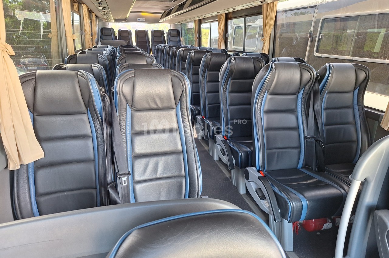 Intérieur d'un autocar avec des rangées de sièges en cuir noir, vides, et des rideaux beige aux fenêtres.