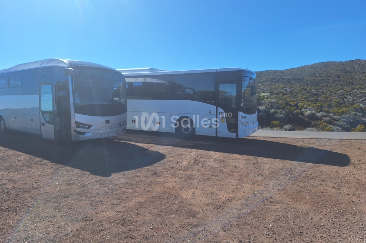 Deux bus blancs stationnés sur un terrain en terre, avec un paysage de collines et de végétation en arrière-plan.