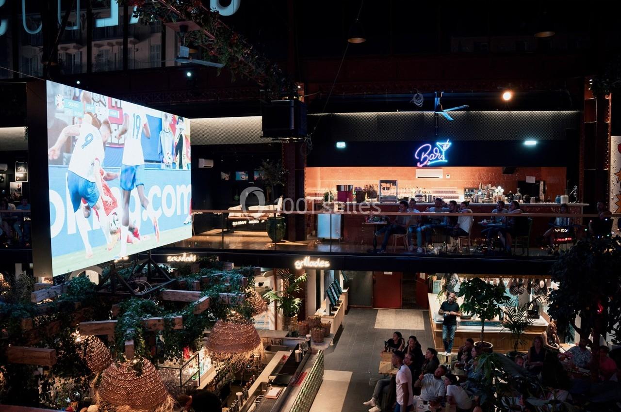 Grand écran diffusant un match de football dans un espace intérieur animé avec bar et spectateurs.