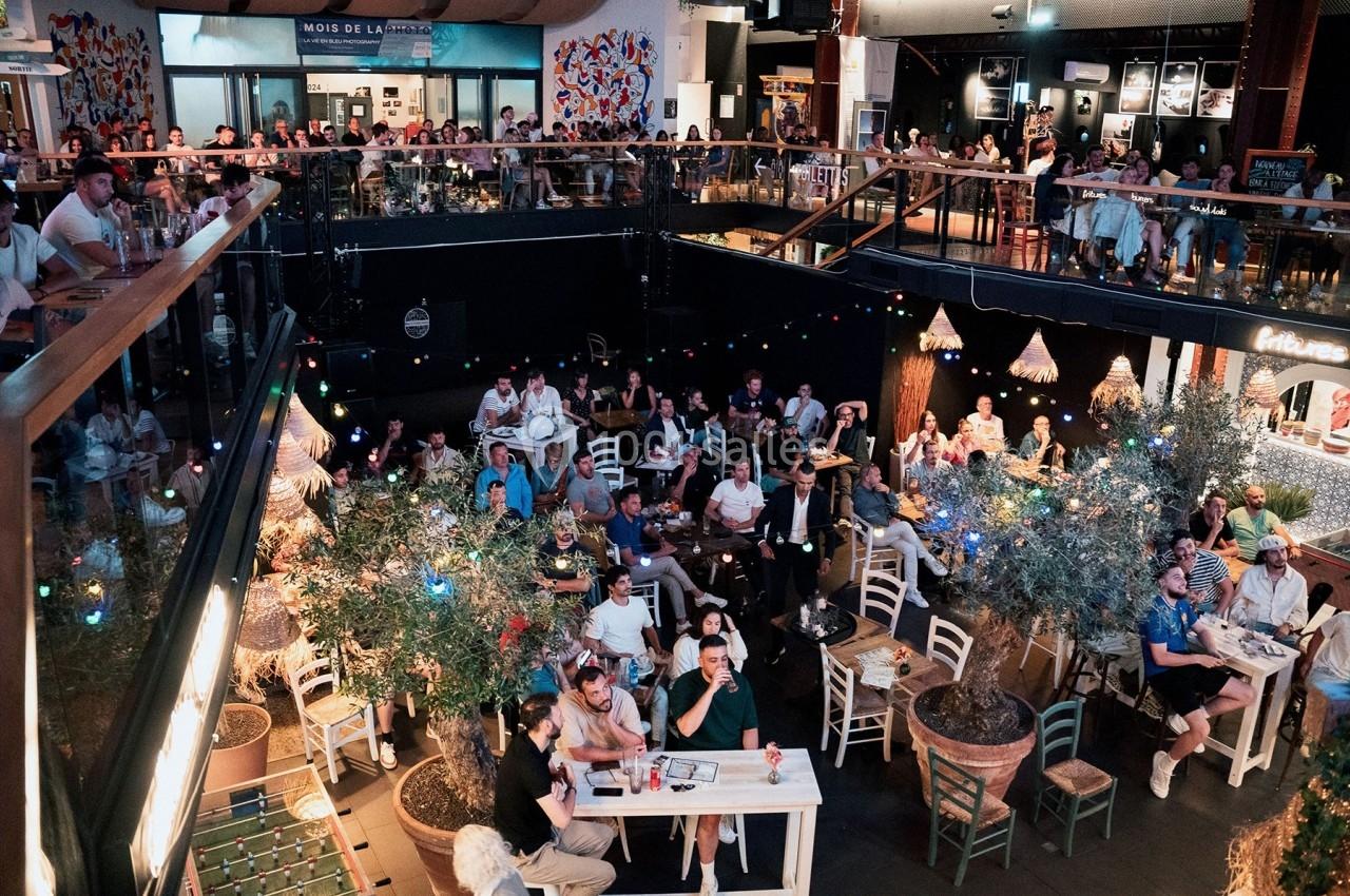 Vue d'une salle animée avec des tables, des chaises et un public assistant à un événement dans un espace intérieur décoré.