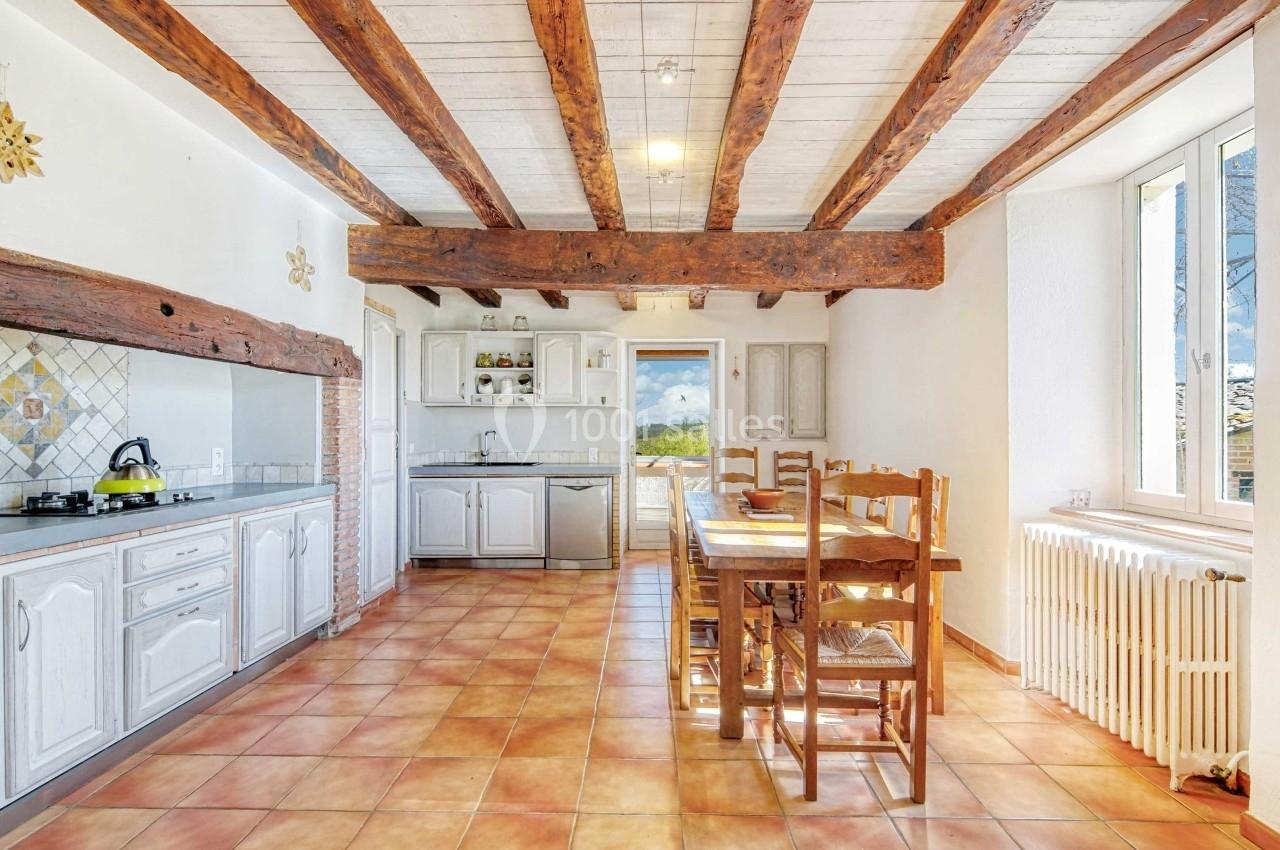Cuisine lumineuse avec poutres apparentes, carrelage au sol, meubles blancs, table en bois et vue sur l'extérieur.