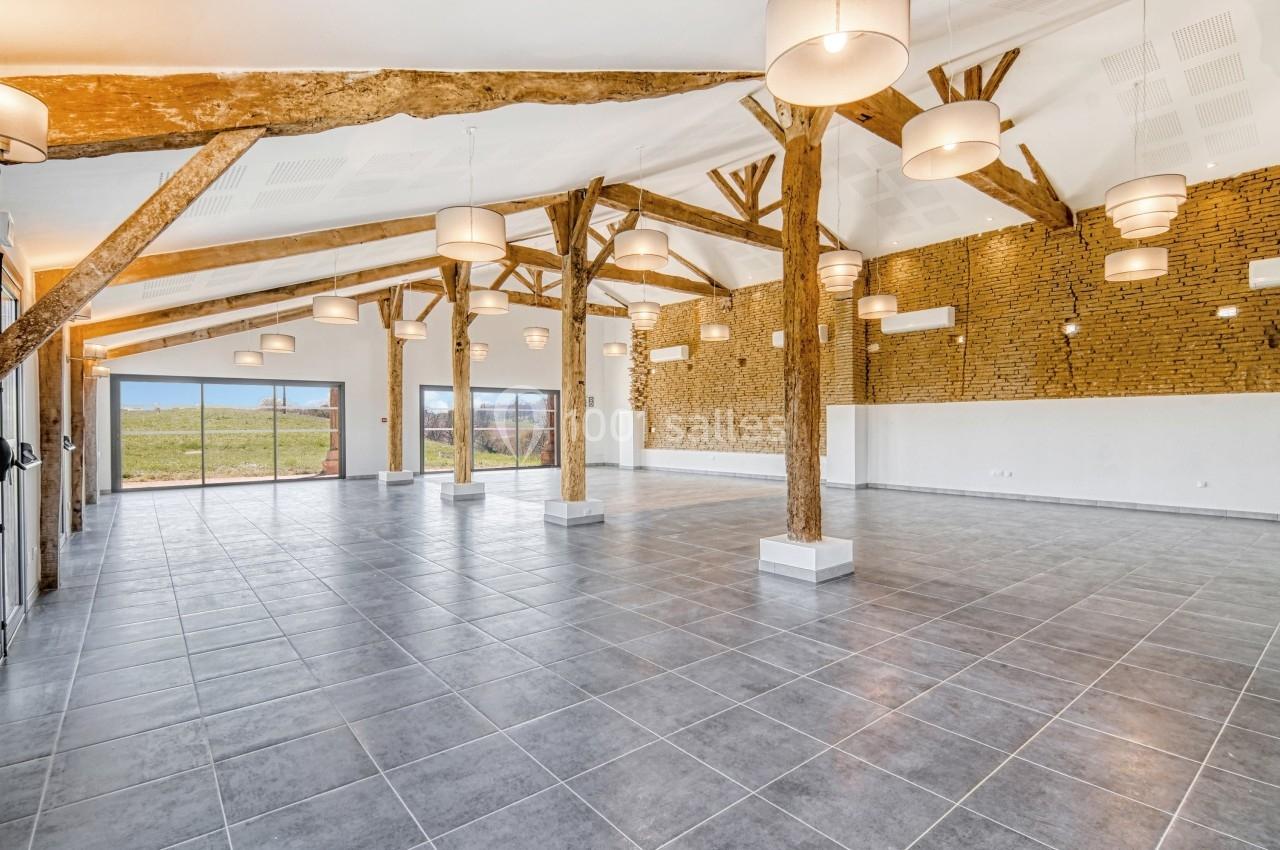 Salle spacieuse avec poutres en bois, murs en briques et carrelage gris, éclairée par des suspensions modernes.