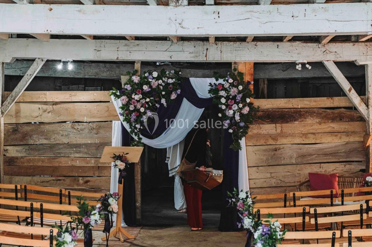 Décoration florale avec arche et drapés dans une grange aménagée pour un événement, rangées de chaises en bois au premier…