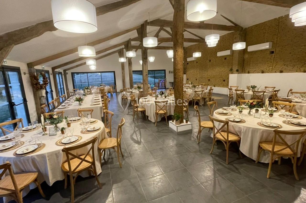 Salle de réception lumineuse avec tables rondes dressées, chaises en bois et poutres apparentes.