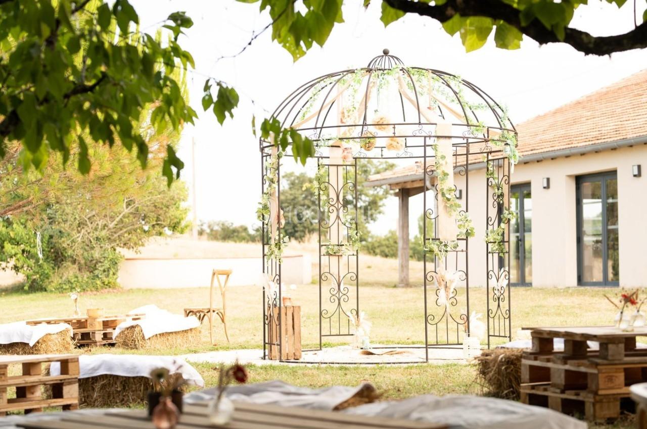Tonnelle en fer forgé décorée de fleurs, entourée de bottes de paille et de tables en bois dans un jardin ensoleillé.