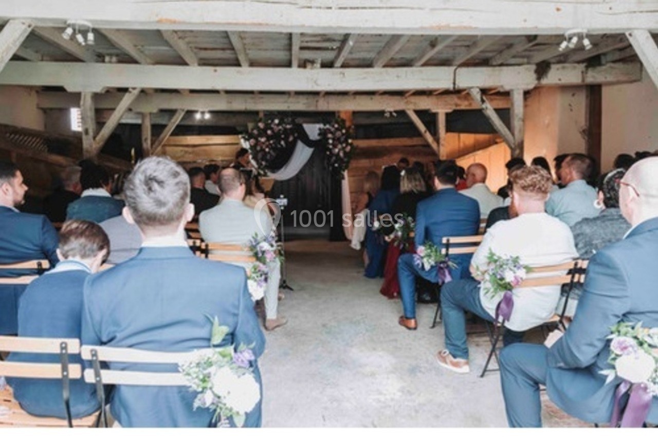 Invités assis dans une grange décorée pour une cérémonie, avec des fleurs attachées aux chaises et une arche florale au fond.
