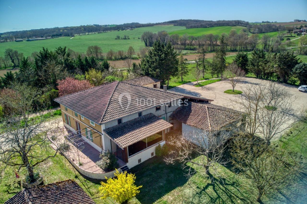 Vue aérienne d'une maison avec toit en tuiles, entourée d'arbres et de champs verdoyants sous un ciel dégagé.