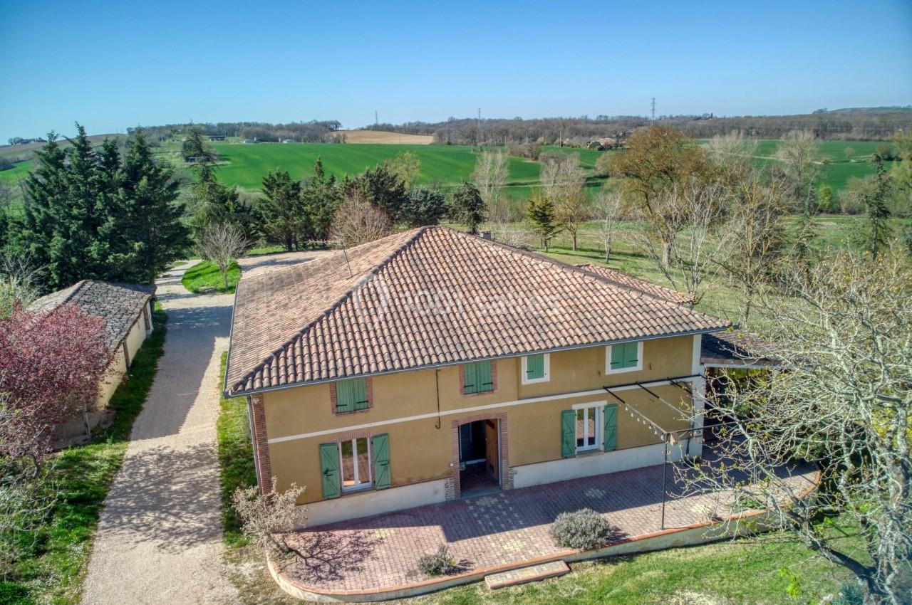 Maison à deux étages avec toit en tuiles, entourée de jardins et de champs verdoyants sous un ciel dégagé.