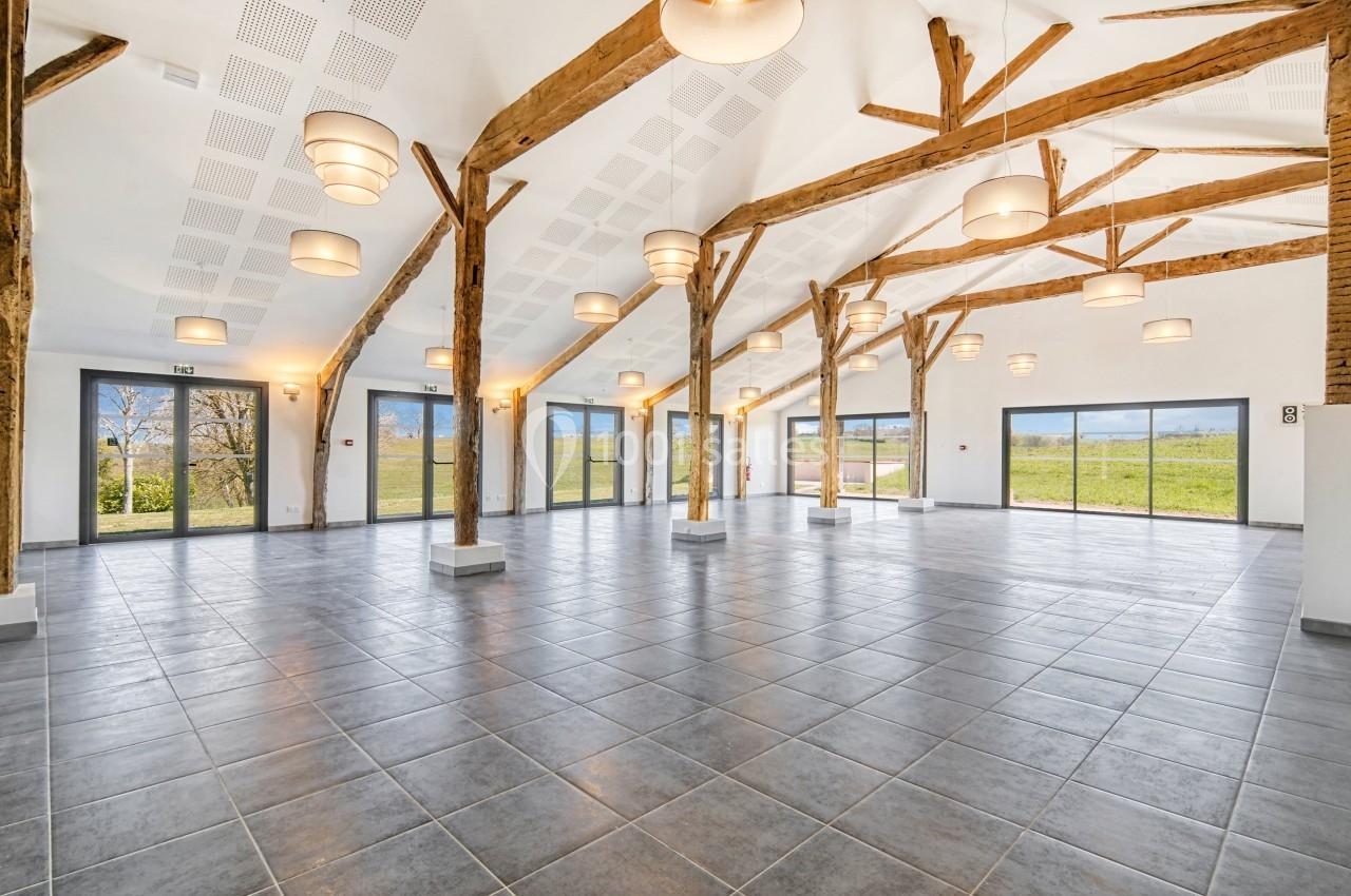 Grande salle lumineuse avec poutres en bois apparentes, carrelage gris et baies vitrées donnant sur un paysage verdoyant.