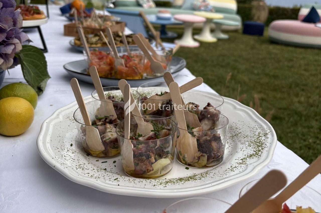 Assortiment de verrines de poulpe mariné avec des cuillères en bois, disposées sur une table en extérieur.