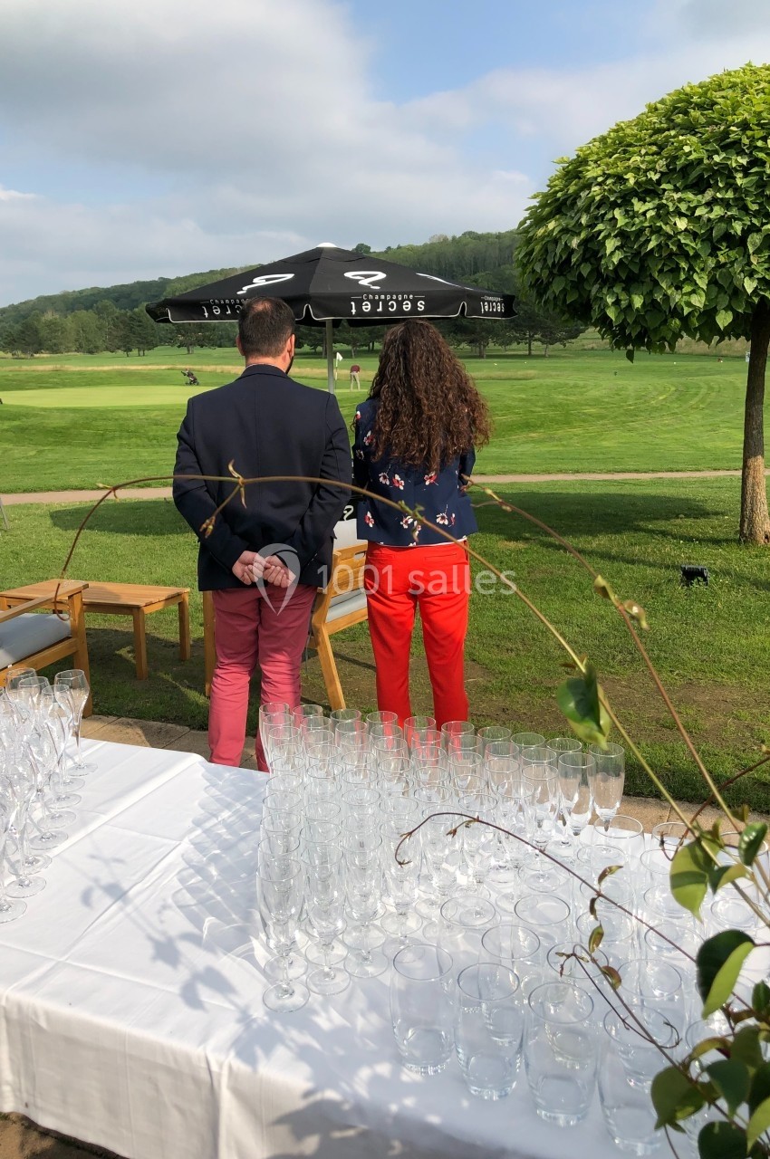Deux personnes de dos regardant un terrain de golf, avec une table dressée de verres au premier plan.
