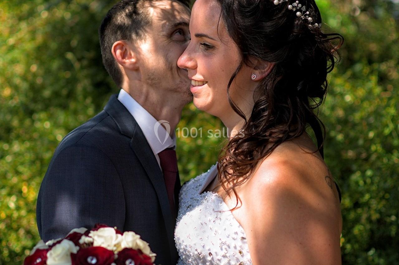Un couple en tenue de mariage pose dans un jardin, la mariée tenant un bouquet de roses rouges et blanches.