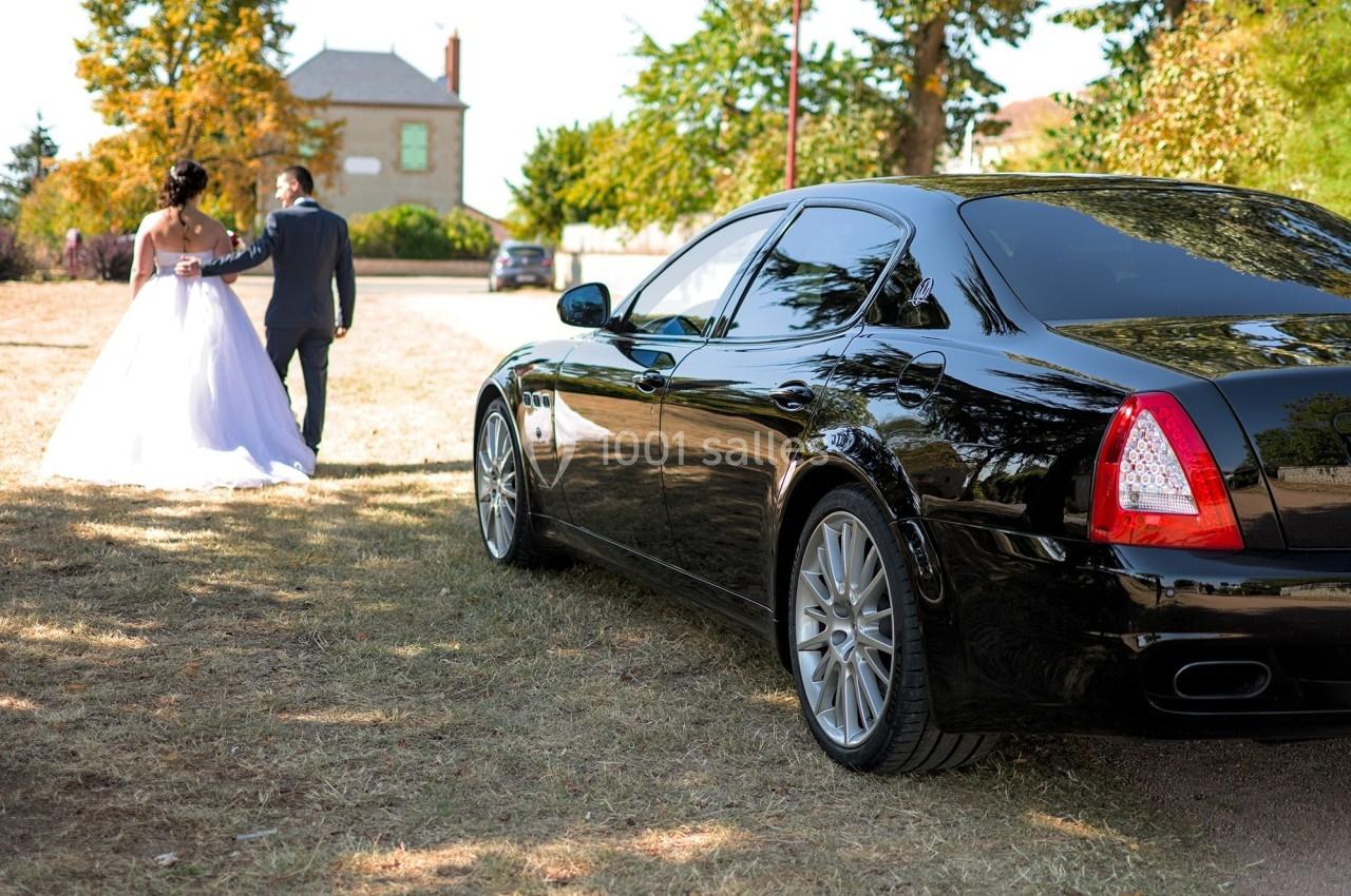 Voiture noire garée sur un chemin herbeux, avec un couple en tenue de mariage marchant au loin.
