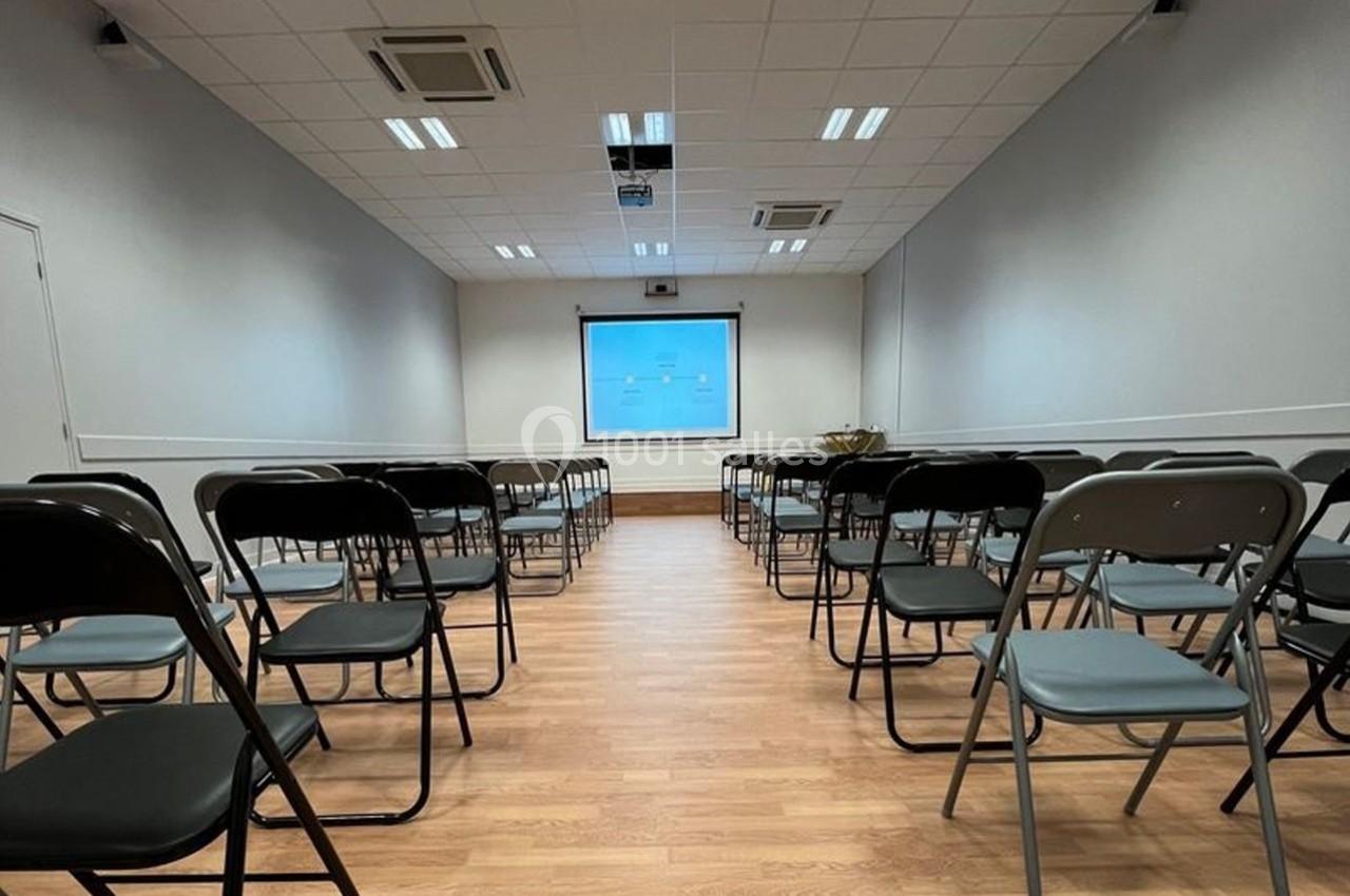 Salle de conférence vide avec des chaises pliantes alignées face à un écran de projection allumé.