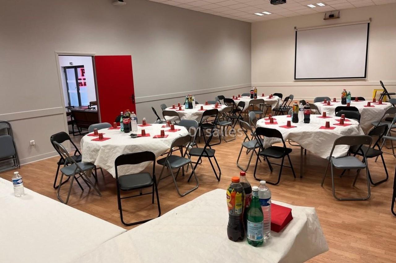 Salle aménagée avec des tables rondes couvertes de nappes blanches, décorées de serviettes rouges et de boissons.