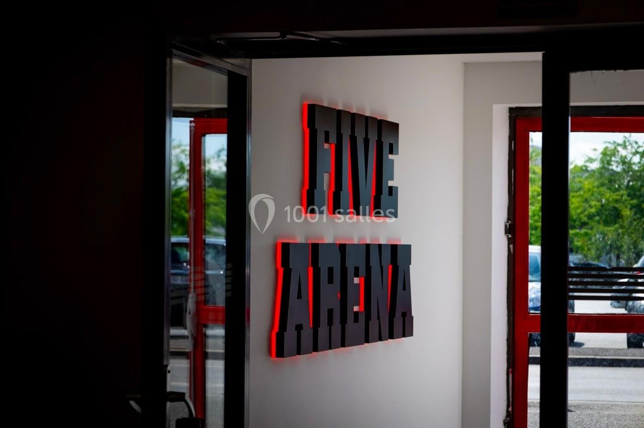 Enseigne murale lumineuse ’FIVE ARENA’ en lettres rouges et noires, visible à travers une porte ouverte.