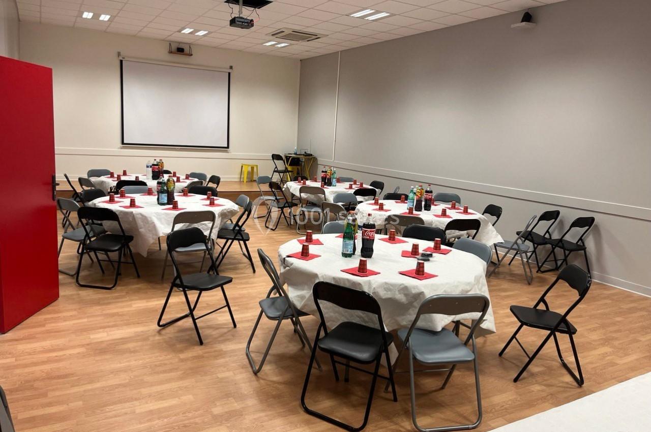 Salle aménagée avec des tables rondes dressées, nappes blanches, chaises pliantes noires et écran de projection en fond.