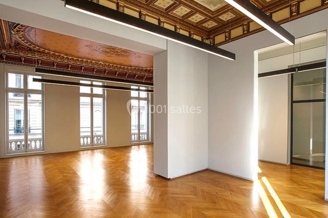 Salle lumineuse avec parquet en bois, plafond décoré et grandes fenêtres donnant sur l'extérieur.