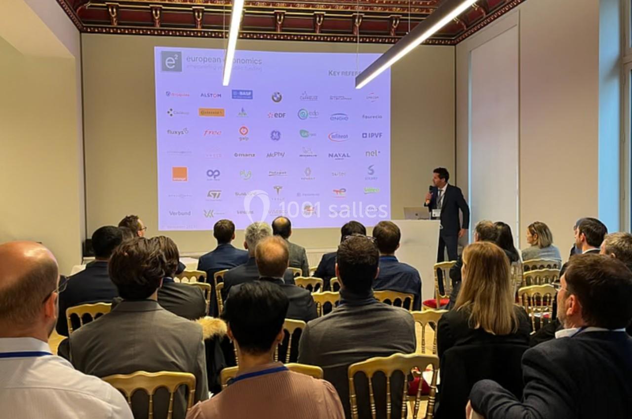 Un conférencier présente devant un public assis, avec un écran affichant des logos d'entreprises en arrière-plan.