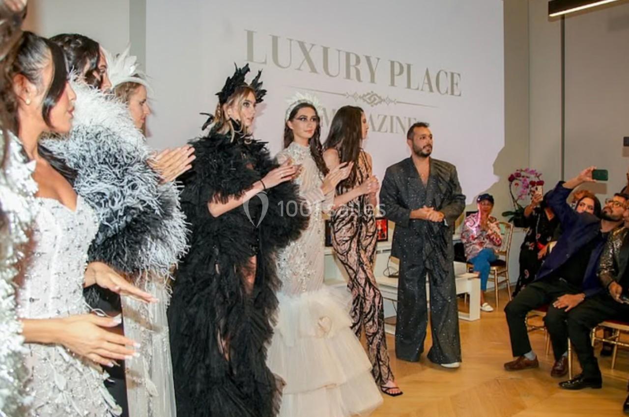 Un groupe de mannequins en tenues élégantes défile sur une scène, observé par un public dans une salle lumineuse.