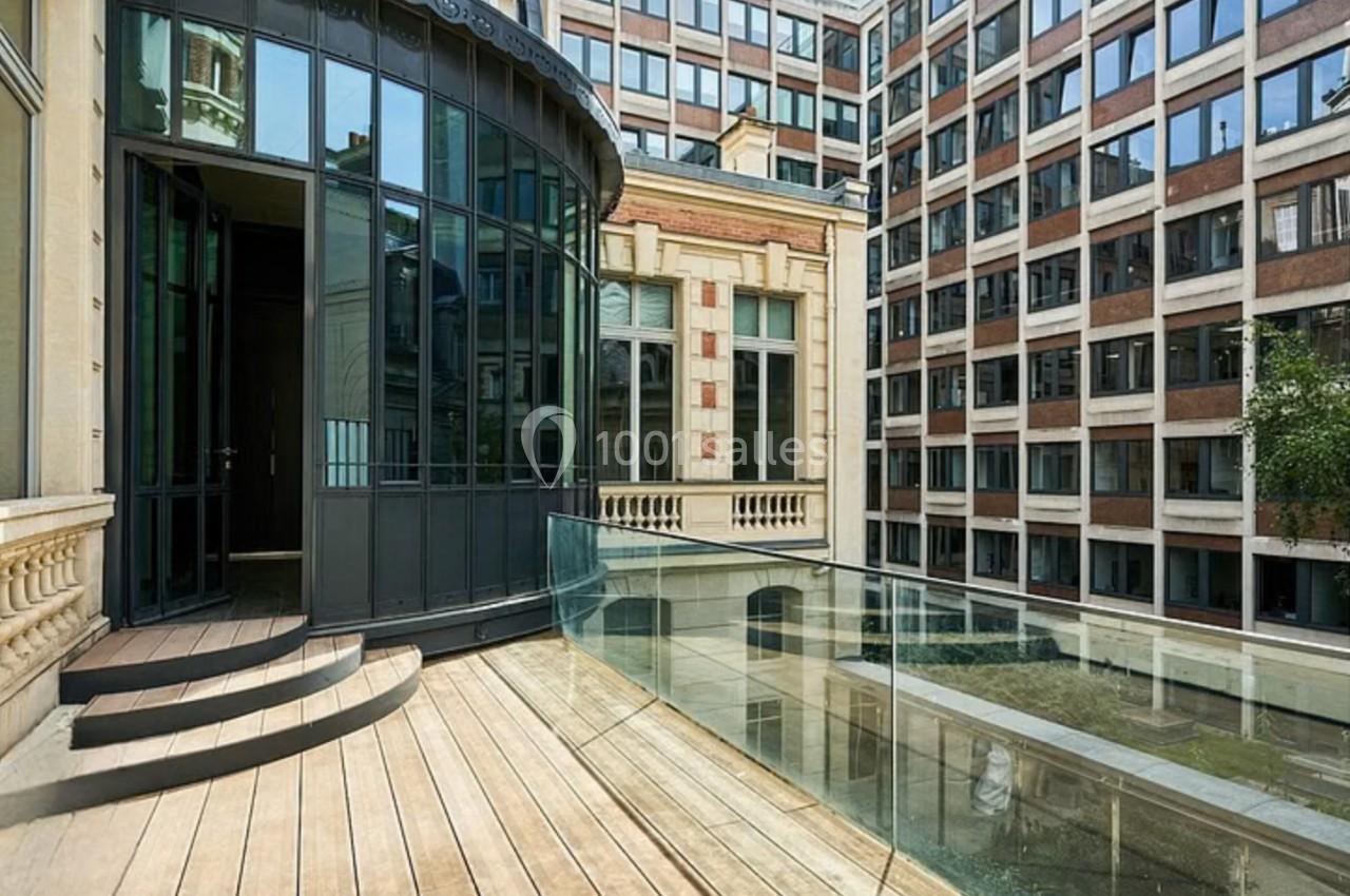 Terrasse en bois avec garde-corps en verre, donnant sur une cour intérieure entourée de bâtiments modernes.