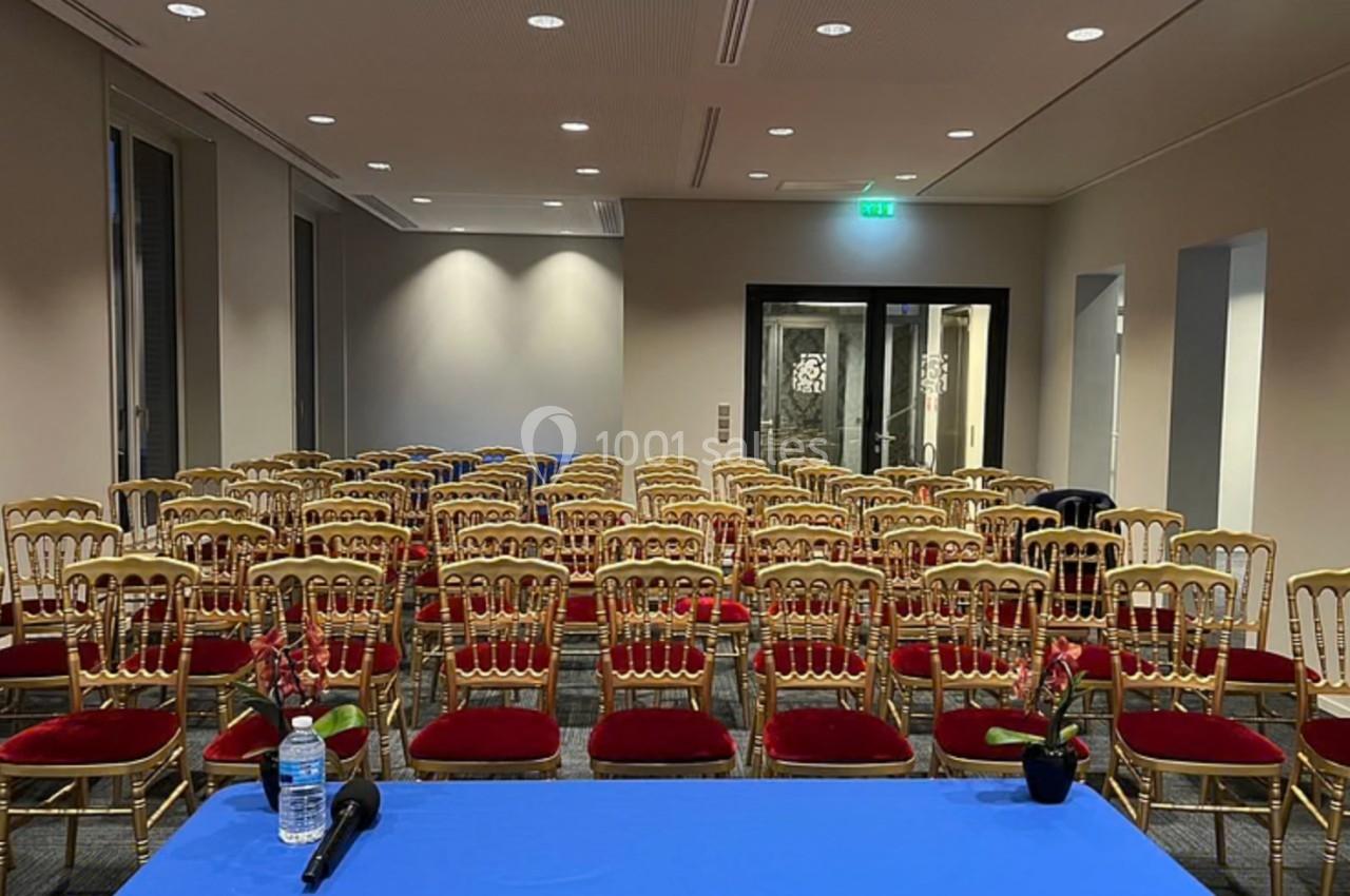 Salle de conférence avec des rangées de chaises dorées et rouges, vue depuis une table avec un micro et une bouteille d'eau.