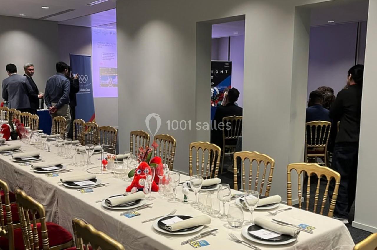 Salle de réception avec tables dressées, chaises dorées et quelques personnes discutant près d'un écran de présentation.