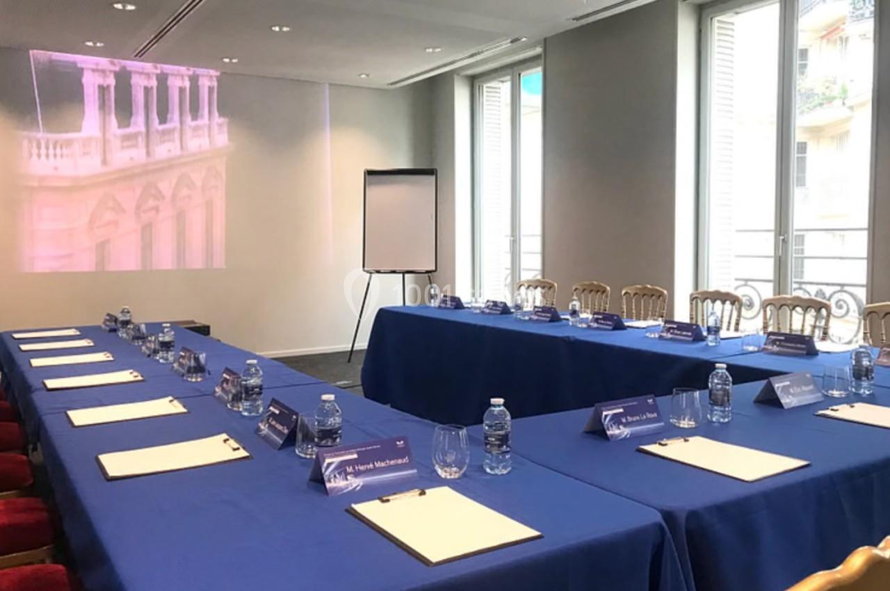Salle de réunion avec tables couvertes de nappes bleues, chaises alignées, bloc-notes, bouteilles d'eau et un tableau blanc.