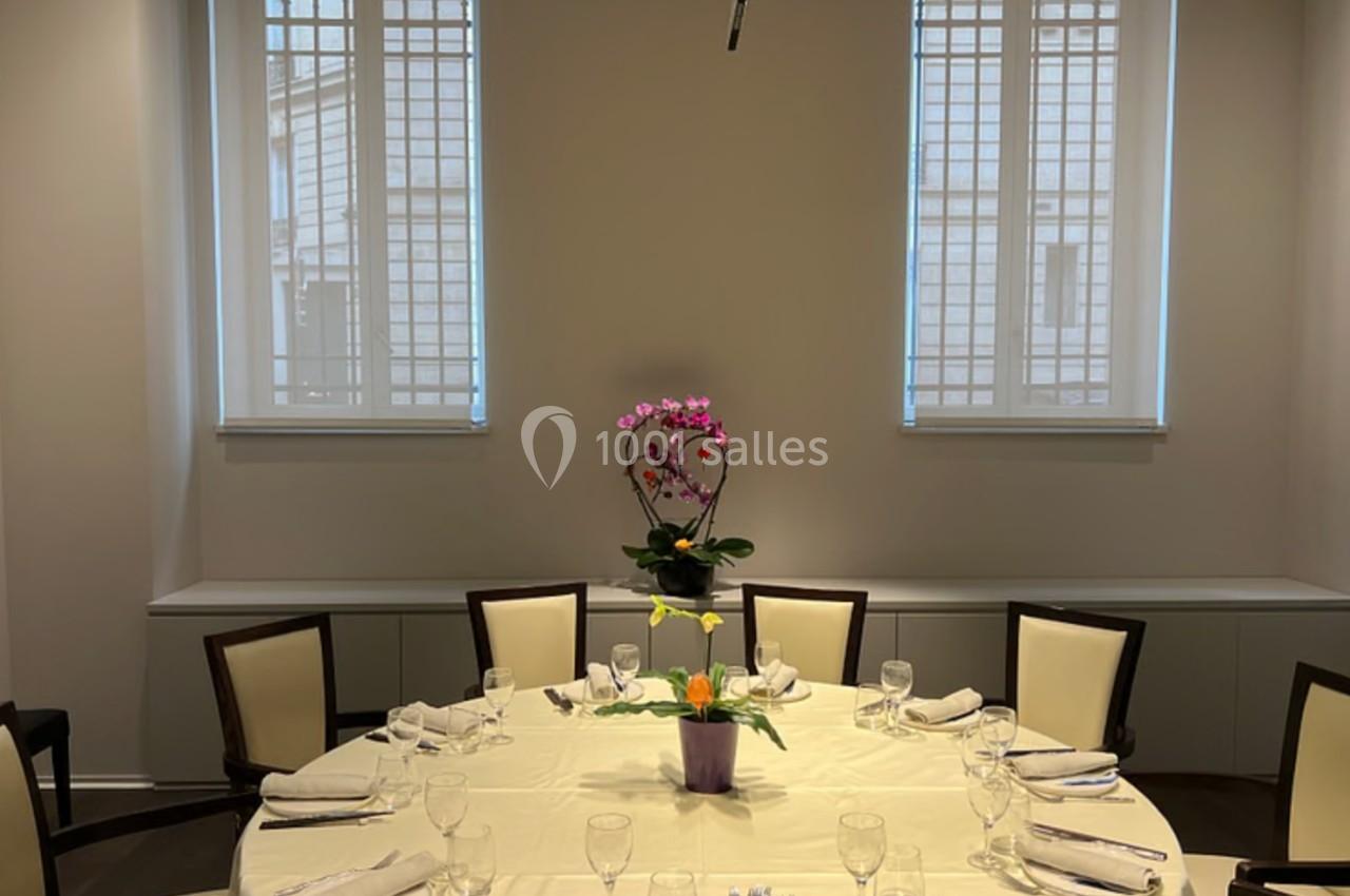 Table ronde dressée avec nappes blanches, verres et serviettes, décorée d'une plante au centre, dans une salle lumineuse.