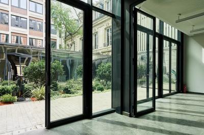 Vue intérieure d'une pièce lumineuse avec de grandes baies vitrées donnant sur un jardin et des bâtiments voisins.
