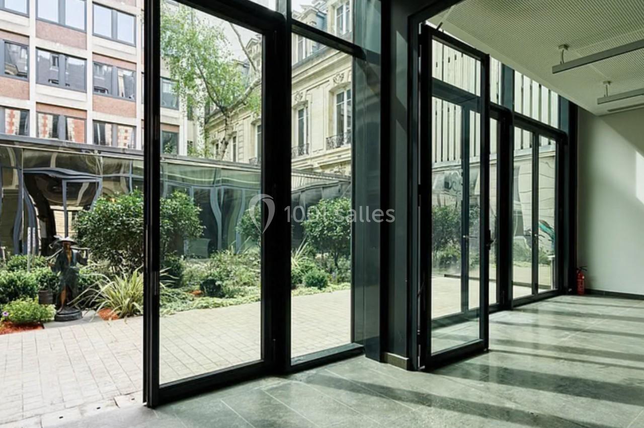 Vue intérieure d'une pièce lumineuse avec de grandes baies vitrées donnant sur un jardin et des bâtiments voisins.