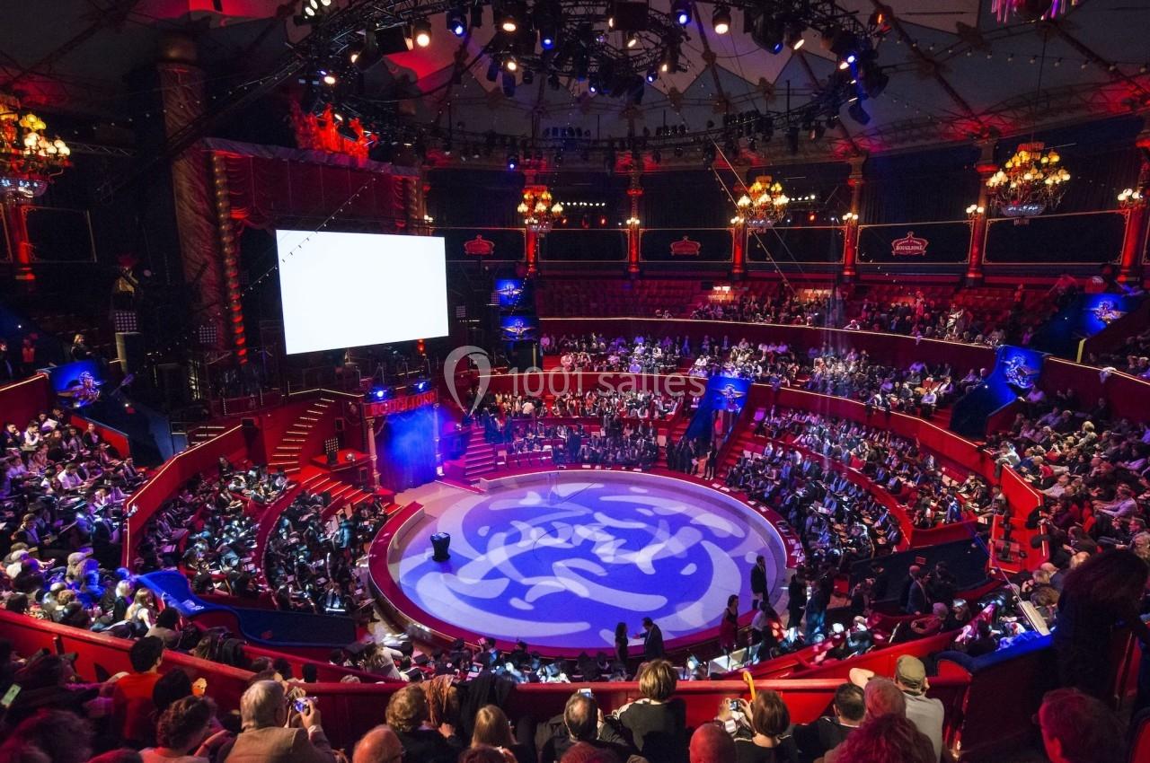 Vue intérieure d'un cirque avec une piste centrale illuminée, entourée de gradins remplis de spectateurs.