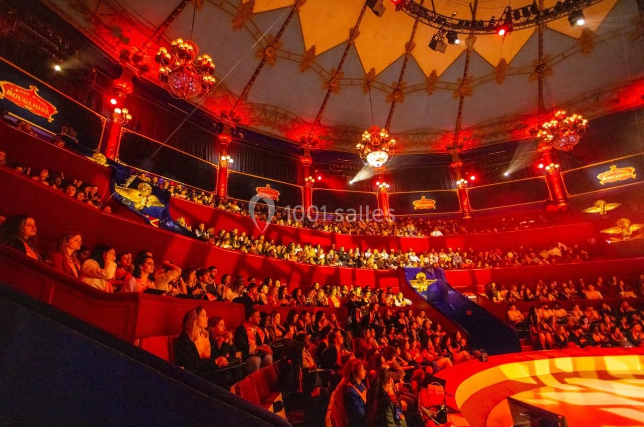 Public assis dans un chapiteau de cirque illuminé de lumières rouges, avec des décorations et des gradins remplis.