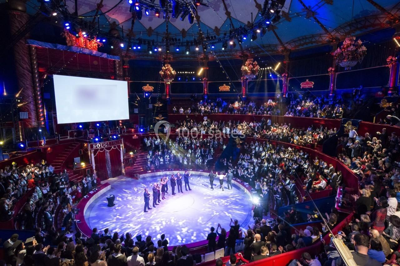 Vue d'une scène circulaire dans un théâtre bondé, éclairée par des projecteurs, avec des artistes alignés au centre.