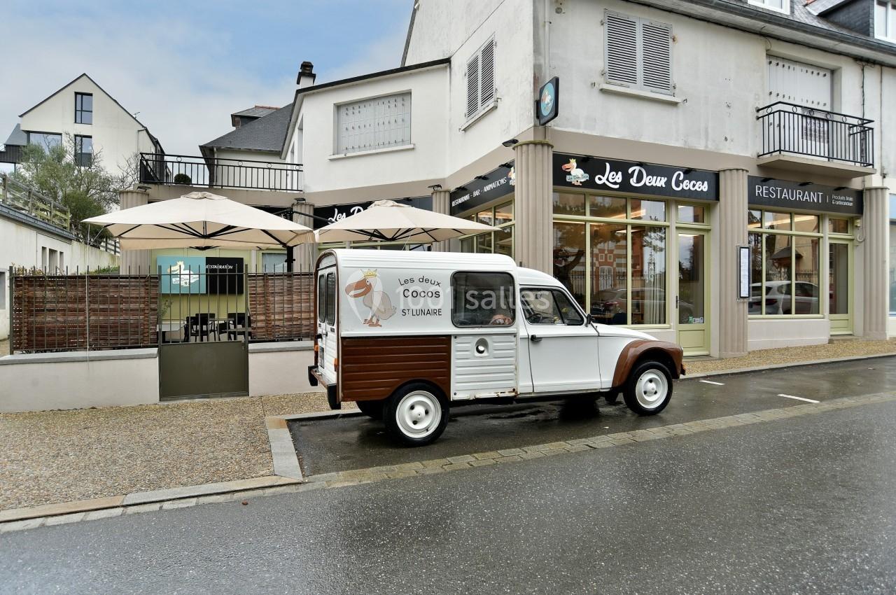Une voiture vintage stationnée devant un restaurant avec terrasse et parasols, dans une rue pavée.