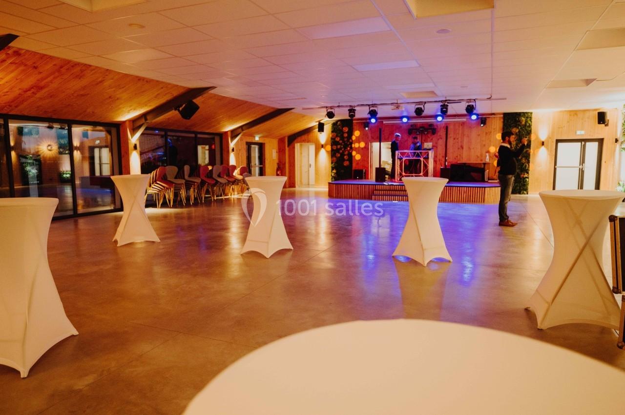 Salle de réception spacieuse avec tables hautes blanches, éclairage coloré et scène équipée pour des événements.