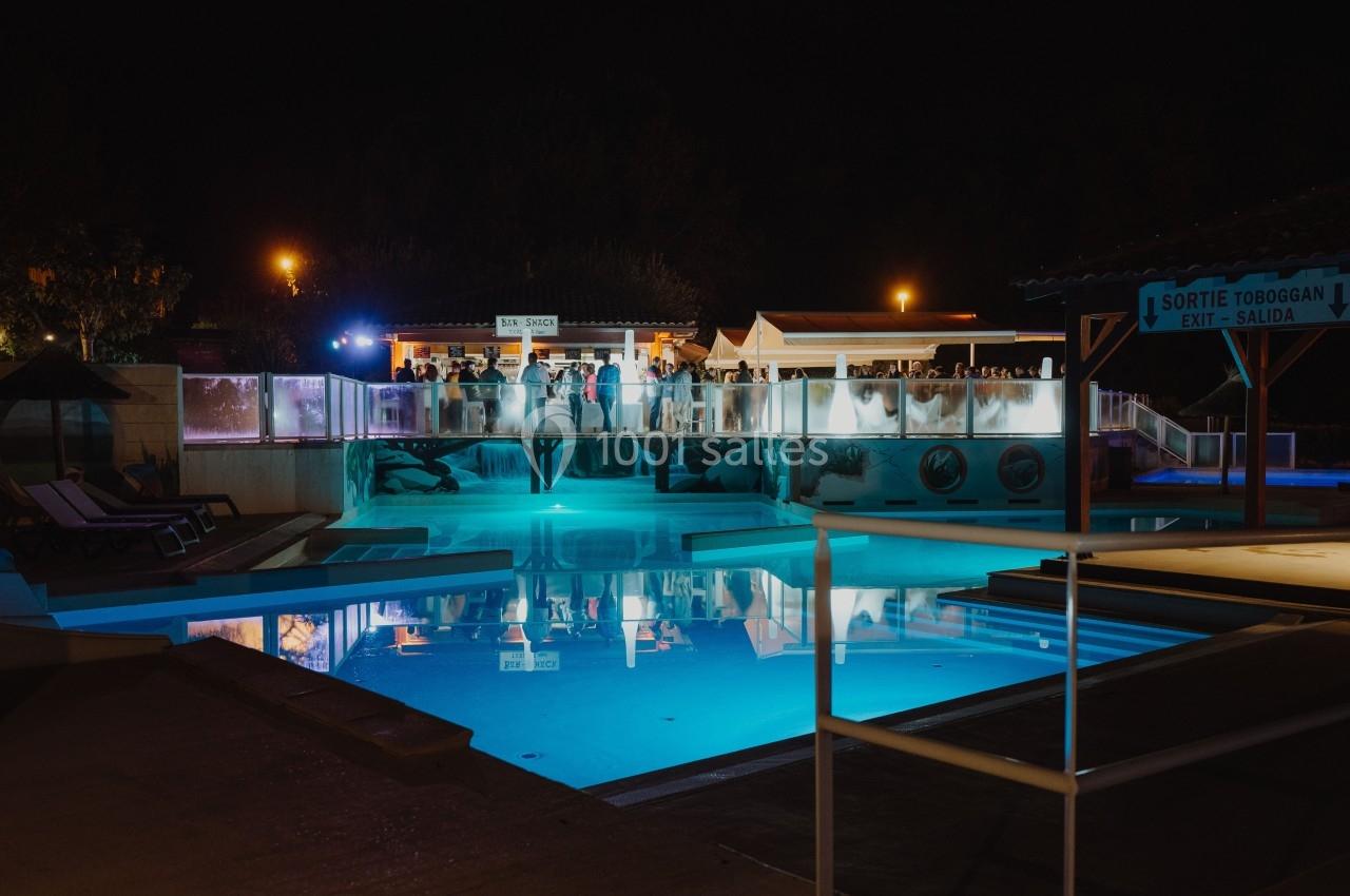Vue nocturne d'une piscine éclairée avec une terrasse animée en arrière-plan et des chaises longues disposées autour.