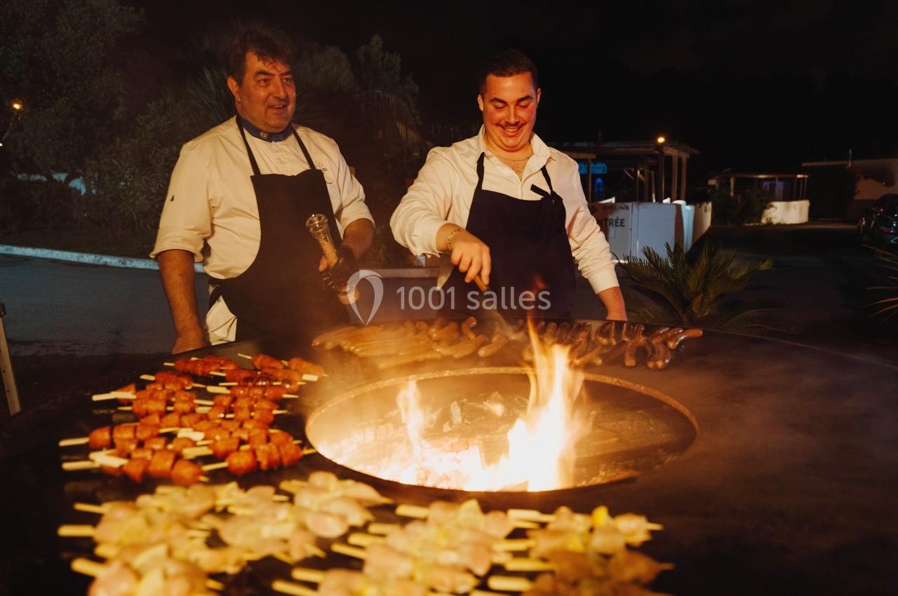 Deux cuisiniers en tablier préparent des brochettes et des saucisses sur un barbecue circulaire en plein air, de nuit.