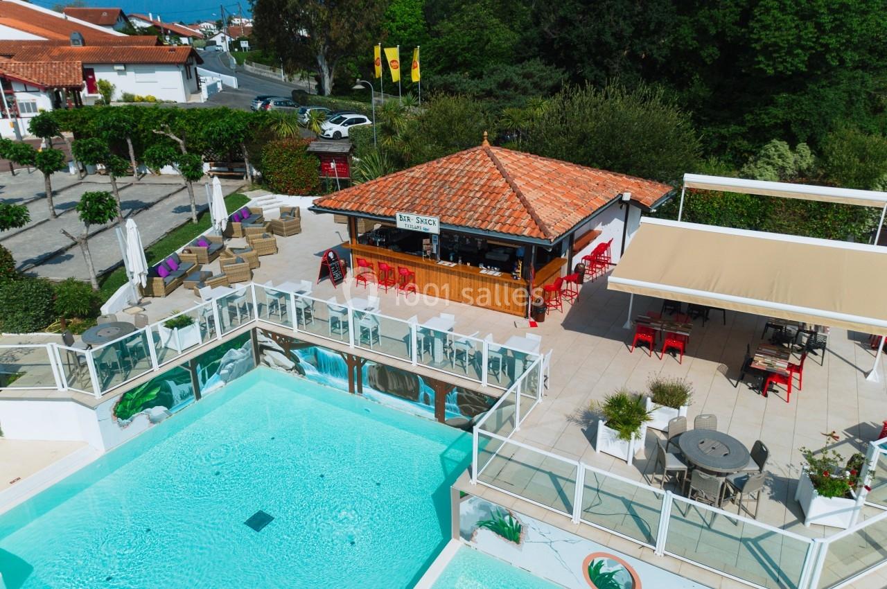 Vue aérienne d'une terrasse avec piscine, bar en toit rouge, tables et chaises, entourée de verdure et bâtiments.