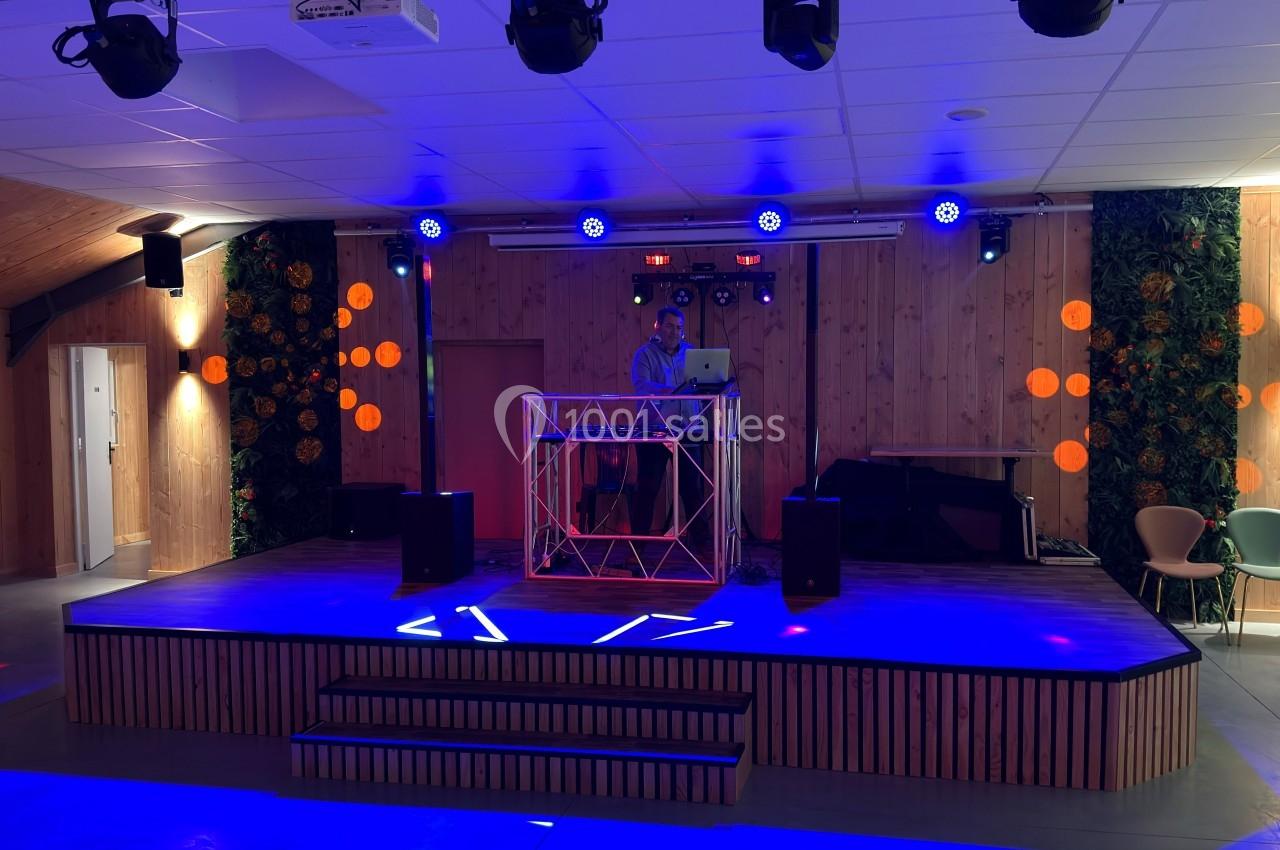 Un DJ mixe sur une scène éclairée par des lumières colorées dans une salle avec des murs en bois et des décorations…