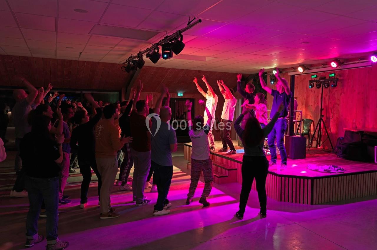Groupe de personnes dansant et applaudissant devant une scène éclairée par des lumières colorées dans une salle intérieure.