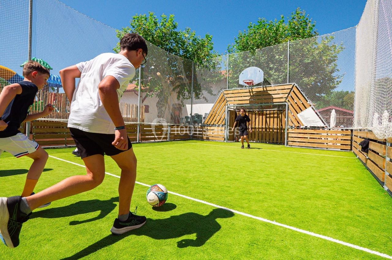 Des enfants jouent au football sur un terrain extérieur clôturé avec un panier de basketball en arrière-plan.