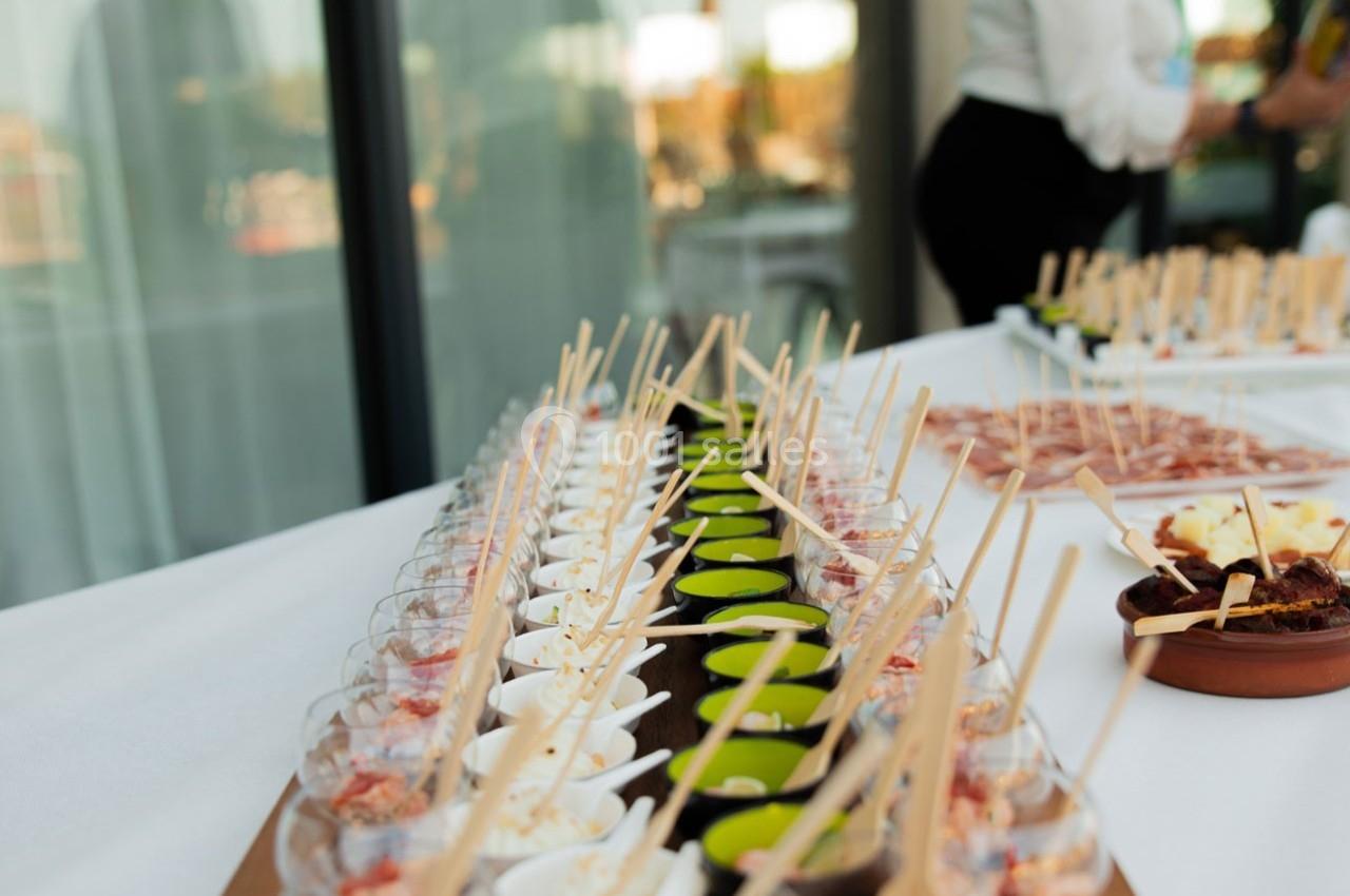Buffet avec verrines colorées et brochettes disposées sur une table blanche, en extérieur près d'une baie vitrée.