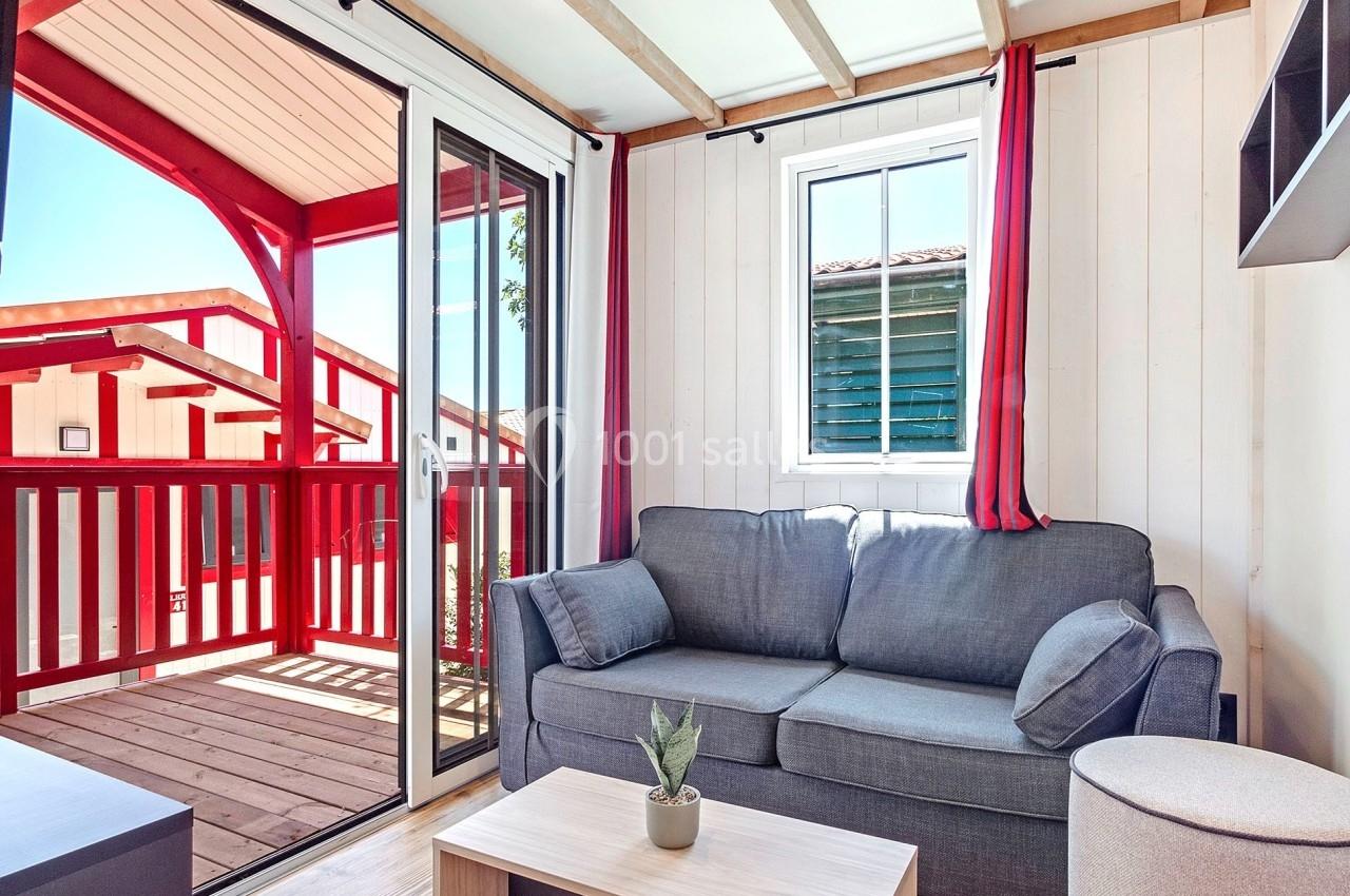 Salon lumineux avec canapé gris, table basse en bois clair, et accès à une terrasse en bois rouge via une baie vitrée.