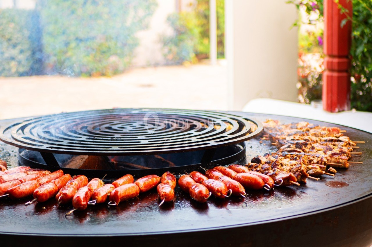 Saucisses et brochettes de viande grillant sur un barbecue circulaire en plein air.
